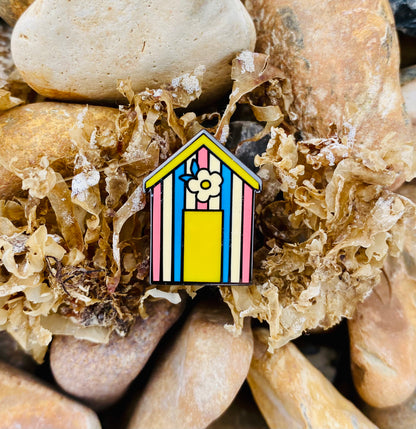 Cute candy stripe beach hut pin badge. The beach hut has pink, blue, yellow and cream stripes and features a yellow door and retro flower above. The photo was taken on the beach pebbles.