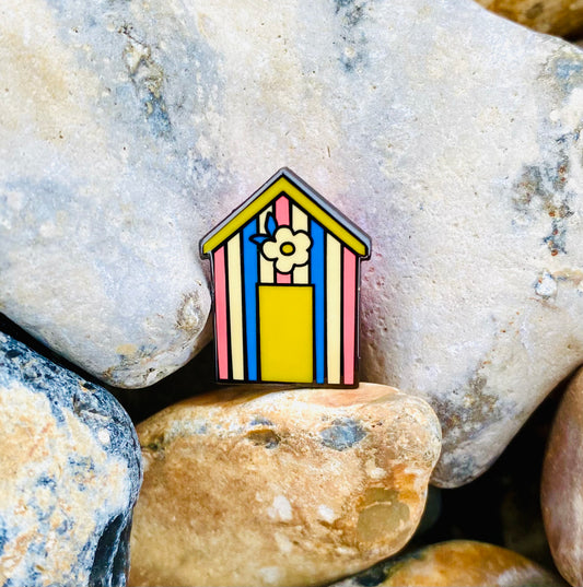 Cute candy stripe beach hut pin badge. The beach hut has pink, blue, yellow and cream stripes and features a yellow door and retro flower above. The photo was taken on the beach pebbles.